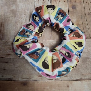 May include: A white scrunchie with a repeating pattern of colorful squares featuring portraits of men. The squares are outlined in various colors, including pink, blue, yellow, and orange. The scrunchie is made of a soft fabric.