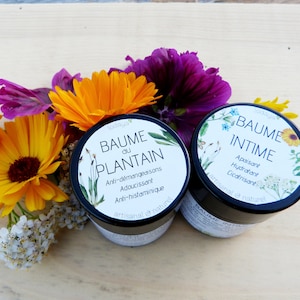 Puede incluir: Dos pequeños frascos de bálsamo natural con diseños florales en las etiquetas. El frasco de la izquierda está etiquetado como "Baume du Plantain" y enumera sus beneficios como "Anti-picazón, Suavizante, Antihistamínico". El frasco de la derecha está etiquetado como "Baume Intime" y enumera sus beneficios como "Calmante, Hidratante, Cicatrizante".