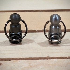 Op de afbeelding: Twee metalen sleutelhangers met een ronde bal aan de bovenkant en een ring aan de onderkant. De sleutelhanger aan de linkerkant is zwart en de sleutelhanger aan de rechterkant is zilver.