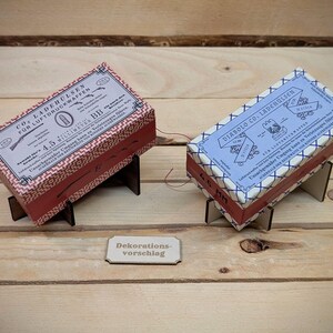 Pode incluir: Duas pequenas caixas com tampas decorativas, uma vermelha com padrões geométricos brancos e pretos e a outra azul com padrões geométricos brancos e pretos. As caixas estão em pequenos suportes de madeira. A caixa vermelha tem o texto "CO. LADENBURGER FOR LOTOF SWIFTER 4.5 FILL. MET. DB" na tampa. A caixa azul tem o texto "DIABOLO CO. LADENBURGER" na tampa. Um pequeno sinal de madeira com o texto "Dekorationsvorschlag" está em primeiro plano.