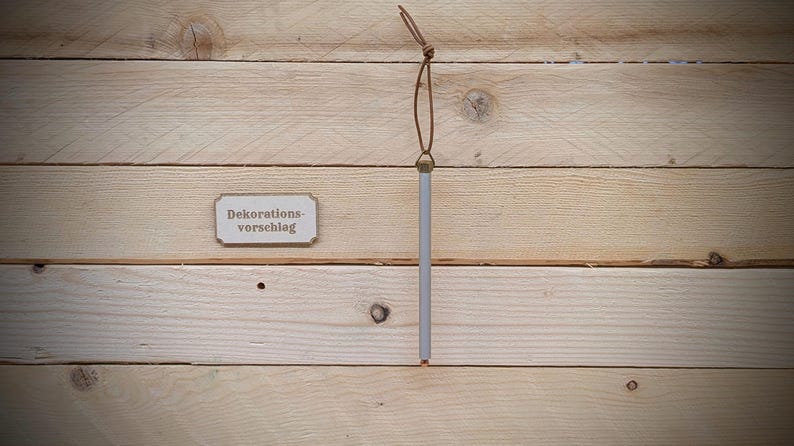 May include: A wooden sign with the text "Dekorations-vorschlag" hanging from a brown leather cord on a light wood background.
