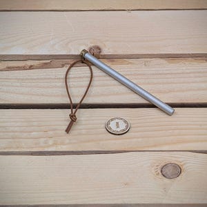 May include: A silver metal fire starter with a brown leather cord and a small round metal disc. The fire starter is on a wooden surface.
