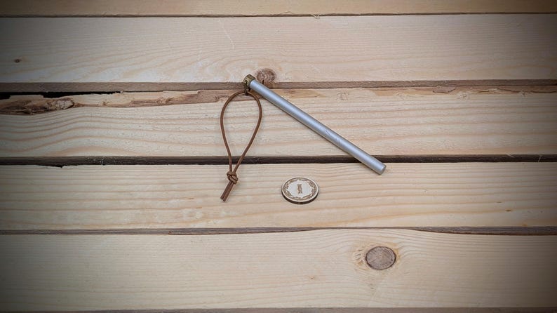 May include: A silver metal fire starter with a brown leather cord and a small round metal disc. The fire starter is on a wooden surface.