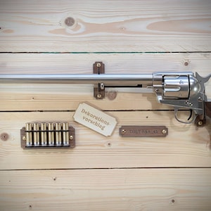 Può includere: Un revolver Colt SAA-45 color argento con impugnatura in legno è montato su uno sfondo di legno chiaro. Sono presenti anche un supporto in legno con proiettili e due piccoli cartelli con testo. I cartelli recitano "Dekorationsvorschlag" e "COLT SAA-45".