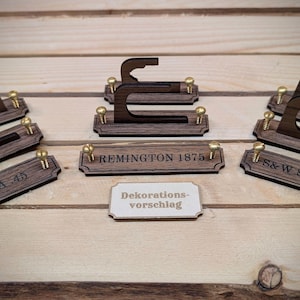 Könnte beinhalten: Dunkelbraune Holz-Waffenständer mit messingfarbenen Schrauben. Die Ständer tragen Gravuren mit den Schriftzügen "COLT SAA 45", "REMINGTON 1875" und "S&W SCHOFIELD". Ein kleines weißes Schild trägt die Aufschrift "Dekorationsvorschlag".