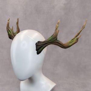May include: A pair of woodland-themed head antlers, featuring a detailed, naturalistic design. The antlers are primarily brown with green and black accents, mounted on a thin, white headband. The headpiece is displayed on a white mannequin head.