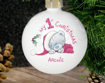 May include: White Christmas bauble with a red ribbon and gold cap. The bauble features a gray bear in a Santa hat and pink outfit, with the text "My 1st Christmas" and the name "Archie".