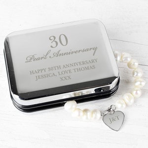 May include: A silver gift box with an engraved message that reads "30 Pearl Anniversary Happy 30th Anniversary Jessica, Love Thomas XXX". A pearl necklace with a heart-shaped charm engraved with "J&T" is placed on top of the box.