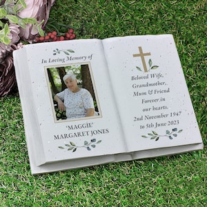 May include: A white ceramic book-shaped memorial with a photo of a woman and the text "In Loving Memory of 'MAGGIE' MARGARET JONES Beloved Wife, Grandmother, Mum & Friend Forever in our hearts. 2nd November 1947 to 5th June 2023"