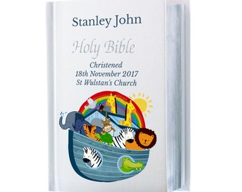 Op de afbeelding: Een witte Heilige Bijbel met de naam "Stanley John" en de woorden "Holy Bible" op de kaft. De kaft heeft een kleurrijke illustratie van dieren in een ark, een regenboog en een zon. De tekst "Christened 18th November 2017 St Wulstan's Church" staat ook op de kaft.