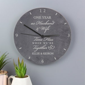 May include: A grey slate clock with black hands and white text that reads "One year as husband & wife Time flies when we're together Ellie & Keiron".