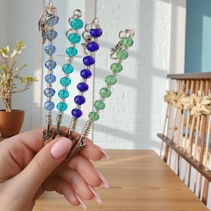 May include: Four beaded clip-on accessories in shades of blue and green. Each clip features a different color scheme of faceted beads strung on a silver wire, with a silver clip at the base. The accessories are held in a hand.