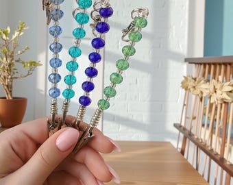 May include: Four beaded clip-on accessories in shades of blue and green. Each clip features a different color scheme of faceted beads strung on a silver wire, with a silver clip at the base. The accessories are held in a hand.