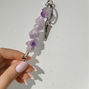 May include: A keychain featuring a strand of amethyst stones in varying shades of purple. The stones are strung together with silver-colored wire and attached to a silver-colored clasp and key ring. The background is white.