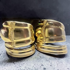 May include: Two gold-colored hoop earrings. Each earring features a thick, curved band with a decorative element on top. The earrings have a shiny, metallic finish and are displayed on a textured surface.