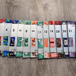 Può includere: Una collezione di quattordici mazzi di carte individuali, ciascuno in una custodia di carta grigio chiaro etichettata con i numeri 3, 4, 4, 5, SIX, 8, NINE, 11, 12, 14, 29, 30, 31, 32 e la parola DEALER. Ogni custodia mostra un motivo colorato unico.