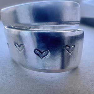 May include: A silver-colored ring with a split design. The lower band features three heart-shaped cutouts. The ring is made of a shiny, metallic material, reflecting light. The ring is a piece of jewelry.
