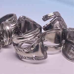 May include: A collection of silver-toned metal rings crafted from repurposed vintage spoons. Each ring features intricate embossed floral and scrollwork patterns along the band, with the curved handle of the spoon forming the circular shape.