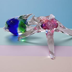 May include: A clear glass sculpture of a fish with a vibrant color palette. The fish features a blue and green head, transitioning to a pink and purple body. The sculpture has a detailed fin and tail design, with a shimmering effect.