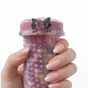 May include: A decorative, cylindrical container covered in small, iridescent beads in shades of purple and clear. The top is a textured pink with a black bow accent. The container is held in a hand with manicured nails.