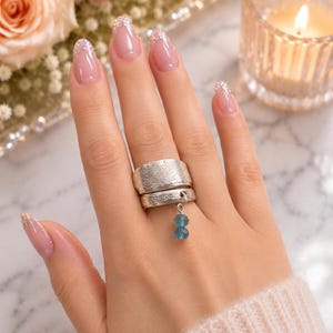 May include: A silver-toned spoon ring featuring an intricate embossed floral pattern across the wide band. A small silver jump ring is attached to the band, holding two stacked, faceted, translucent blue beads that dangle vertically.