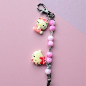 Puede incluir: Un llavero con dos figuras de Hello Kitty, una con un vestido rosa y la otra con un lazo rojo. El llavero tiene un cierre plateado, cuentas rosas y blancas y un clip plateado. El fondo es de color rosa liso.