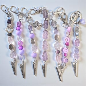 Puede incluir: Una colección de siete cordones con cuentas en tonos morados y plateados. Cada cordón presenta un clip plateado y una variedad de cuentas, incluyendo cuentas facetadas, redondas y metálicas decorativas. Algunos tienen dijes como un árbol o una carita sonriente.