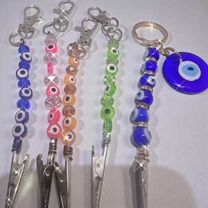 May include: A collection of beaded keychains with silver-tone clips. The keychains feature various colors of beads, including blue, pink, orange, and green, with evil eye designs. One keychain has a blue evil eye charm.