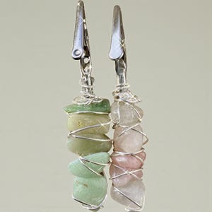 Puede incluir: Dos clips decorativos de metal plateado, cada uno adornado con una pila de piedras preciosas de colores. Uno presenta piedras verdes y amarillas, mientras que el otro muestra piedras rosas y blancas, todo envuelto en alambre plateado.