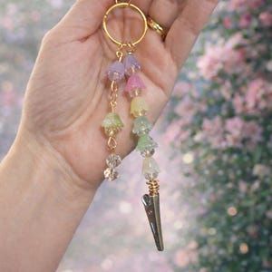 May include: A gold-colored keychain with a clip and a ring. The keychain features a series of pastel-colored flower charms and clear crystal beads. The bottom of the keychain has a gold-colored clip.