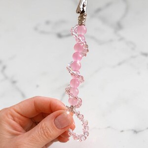 May include: A metal alligator clip attached to a decorative handle featuring a spiral design of matte pink spherical beads and smaller, clear faceted crystal beads. The beads are arranged in a twisted pattern leading to a circular loop at the base.