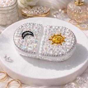Può includere: Un contenitore bianco a forma di pillola, tempestato di strass, con un design a sole e luna. Il contenitore è diviso in due sezioni, una con una luna crescente nera e l'altra con un sole giallo. Il contenitore è circondato da strass e poggia su una superficie bianca.