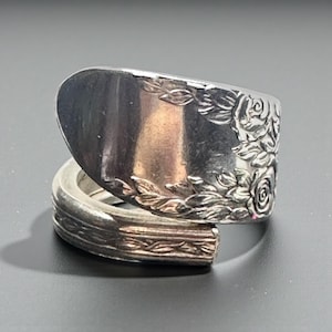 Puede incluir: Un anillo de cuchara plateada con un diseño floral de rosas. El anillo está hecho de una cuchara reutilizada, con la cuchara formando la parte superior del anillo. La banda presenta un patrón floral detallado.