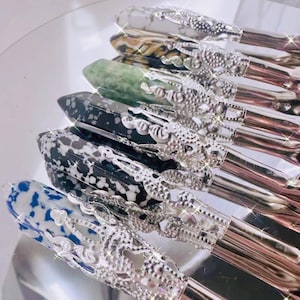 May include: A collection of makeup brushes with decorative silver handles and patterned tops. The tops feature various designs, including white and blue speckled, black and white speckled, and green. The brushes are arranged on a reflective surface.