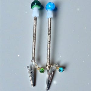 May include: Two silver-toned hair clips with mushroom-shaped toppers. One mushroom is green and white, the other is blue and white. Each clip has a dangling charm with a gemstone, one green and one blue.