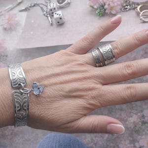 May include: A silver bracelet with an ornate design and a butterfly charm, along with several silver rings, are displayed on a hand. The jewelry is set against a soft, floral background, creating a delicate and feminine aesthetic.