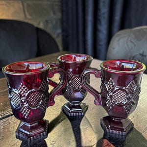 May include: Three ruby red glass mugs featuring a textured diamond and circle pattern on the exterior. Each mug has a sturdy handle and a hexagonal base, crafted from pressed glass with a deep crimson hue and a reflective, polished finish.