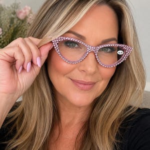 Puede incluir: Gafas de lectura con forma de ojo de gato y montura de color rosa claro, totalmente incrustada con pequeños y brillantes diamantes de imitación rosas. Las lentes son transparentes y llevan una pequeña pegatina circular con la graduación +2,75.