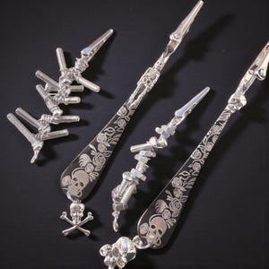 May include: Three silver-toned metal alligator clips featuring decorative handles engraved with floral patterns and skull motifs. One clip has a skeleton figure, another has a skull and crossbones charm, and all are adorned with various metal hardware components.