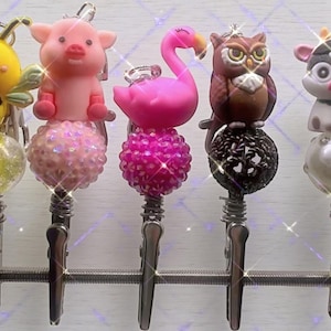 May include: A collection of decorative badge reels featuring animal-themed toppers. The toppers include a pink pig, a pink flamingo, and a brown owl. Each reel has a decorative beaded ball and a silver clip.