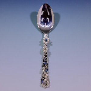 May include: A decorative silver spoon with a floral design. The handle is decorated with white pearls, blue beads, and silver wire. The spoon has a polished, reflective surface.