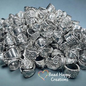May include: A pile of silver rings, each crafted from ornate silverware. The rings feature floral and scroll designs, showcasing a vintage aesthetic. The rings are piled together, reflecting light and creating a textured, metallic display. The text "Bead Happy Creations" is visible.