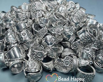 May include: A pile of silver rings, each crafted from ornate silverware. The rings feature floral and scroll designs, showcasing a vintage aesthetic. The rings are piled together, reflecting light and creating a textured, metallic display. The text "Bead Happy Creations" is visible.