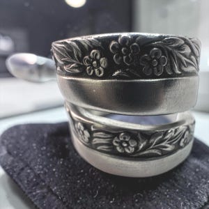 以下が含まれることがあります： 2つの銀色のリングが積み重ねられています。リングの上端には花の模様があります。リングは黒い表面に置かれています。