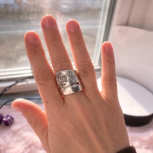 Può includere: Un anello d'argento realizzato con posate riciclate. L'anello presenta un motivo floreale sulla fascia superiore e una fascia liscia sottostante. L'anello è indossato su un dito, mettendo in risalto il suo stile unico e artigianale.