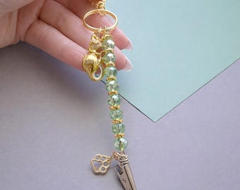 May include: A gold-colored keychain with a cat charm, green beaded accents, and a paw print charm. The keychain also features a silver-colored alligator clip.