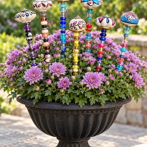 May include: Seven decorative garden stakes featuring ceramic knobs and colorful glass beads in various patterns and hues, inserted into a black fluted urn filled with purple chrysanthemum flowers. Each stake is topped with a unique ceramic finial.