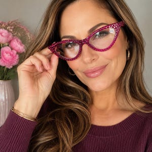 Könnte beinhalten: Pinkfarbene Katzenaugen-Lesebrille mit Strasssteinen. Die Fassungen sind leuchtend magenta, und die Gläser haben eine Vergrößerung von +2,00. Die Brille wird gehalten.