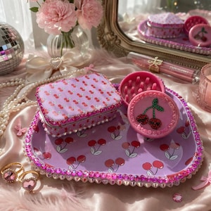 May include: A decorative tray, box, and compacts in a matching cherry design. The tray and box are lavender with red cherries, and the compacts are pink. The tray is trimmed with pink rhinestones. Two heart-shaped rings are in the foreground.
