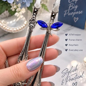 May include: Two silver-toned metal clips featuring decorative embellishments. One clip has a silver flower design, while the other features a dragonfly with blue faceted stones. Both clips have a spring-loaded mechanism and a loop at the base.
